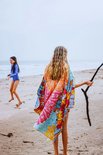 Meisje met lang blond haar, deels bedekt door een kleurrijk strandlaken met mandala print, staat op het strand met de zee op de achtergrond.
