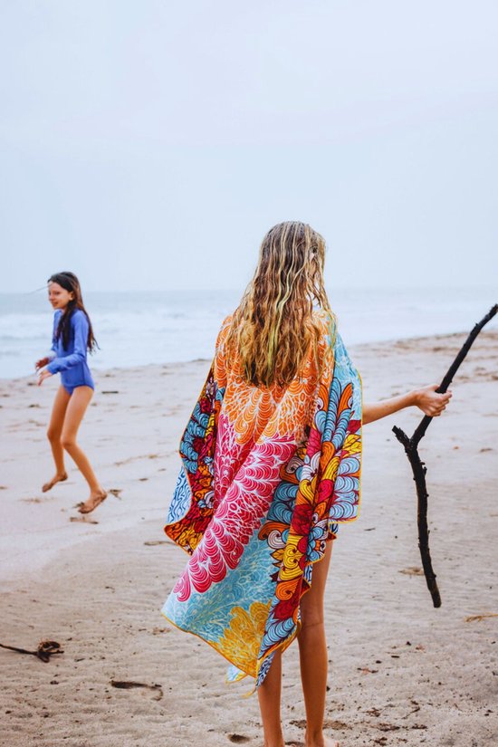 Meisje met lang blond haar, deels bedekt door een kleurrijk strandlaken met mandala print, staat op het strand met de zee op de achtergrond.