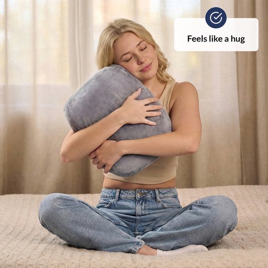 Een vrouw zit op een bed en omhelst een grijs verzwaringskussen.