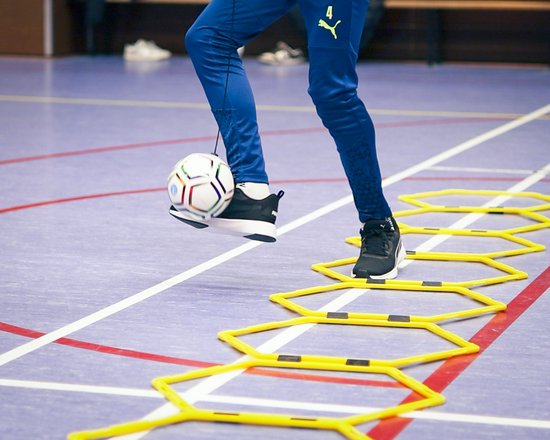 Close-up van benen van een jongen die op een voetbal traint, met gele agility-ringen op de grond.