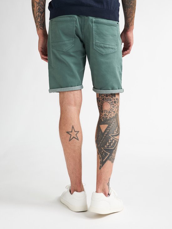 Man met tatoeages op zijn benen, draagt een groene denimshort en witte sneakers.