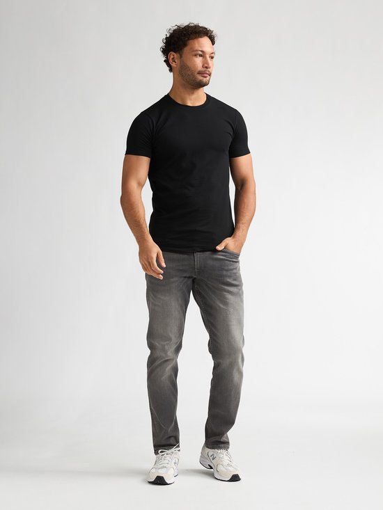 Man met handen in broekzakken, draagt zwart T-shirt met korte mouwen en grijze jeans.