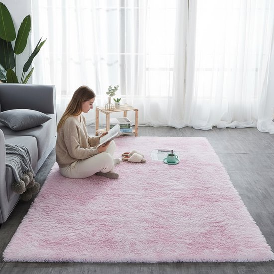Een vrouw zit op een lichtroze, fluffy vloerkleed, lezend in een woonkamer.