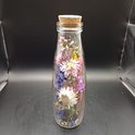 Glazen fles met kurk, gevuld met gedroogde bloemen in roze, paars, wit en blauw.
