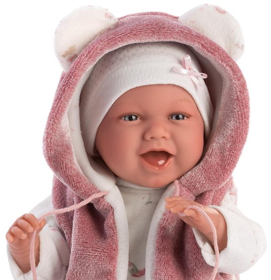 Close-up van lachende babypop Mimi met berenmuts en roze vestje, die speelt met de koordjes.