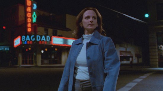 Een vrouw met bruin haar en een blauwe jas loopt 's nachts over straat, voor een theater met neonreclame.