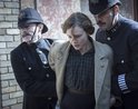 Vrouw in beige jas wordt gearresteerd door twee politieagenten in donkere uniformen en helmen.