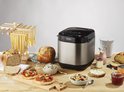 Zilverkleurige broodbakmachine op een houten tafel, omringd door diverse broden, pasta, beleg en ontbijtproducten.