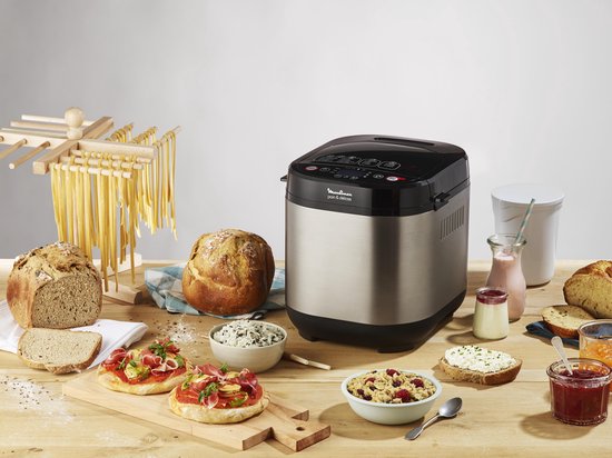 Zilverkleurige broodbakmachine op een houten tafel, omringd door diverse broden, pasta, beleg en ontbijtproducten.