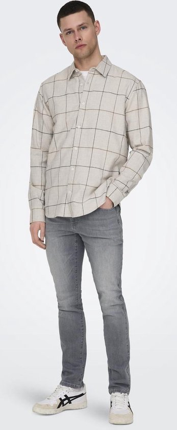 Man in grijze slim fit jeans en geblokt shirt. Witte sneakers. Hand in broekzak.