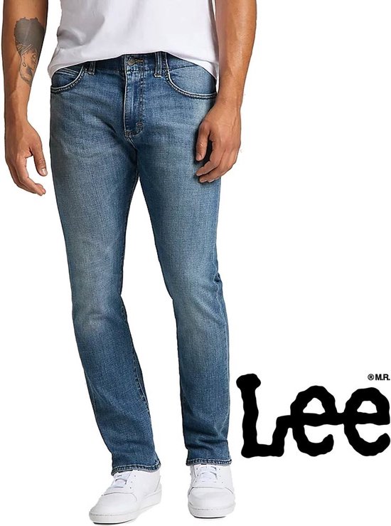 Man in een lichtblauwe jeans, staand met witte sneakers en het Lee-logo op de zijkant.