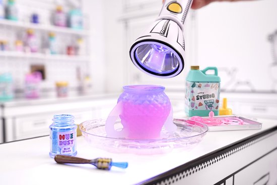 Een miniatuur vaasje dat paars gloeit onder een UV-lamp, met een flesje 'HUE DUST' ernaast.