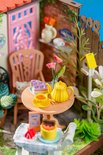Miniatuur tuintafel met gele theepot, gebak, en bloemen. Een katje zit op de stoel.