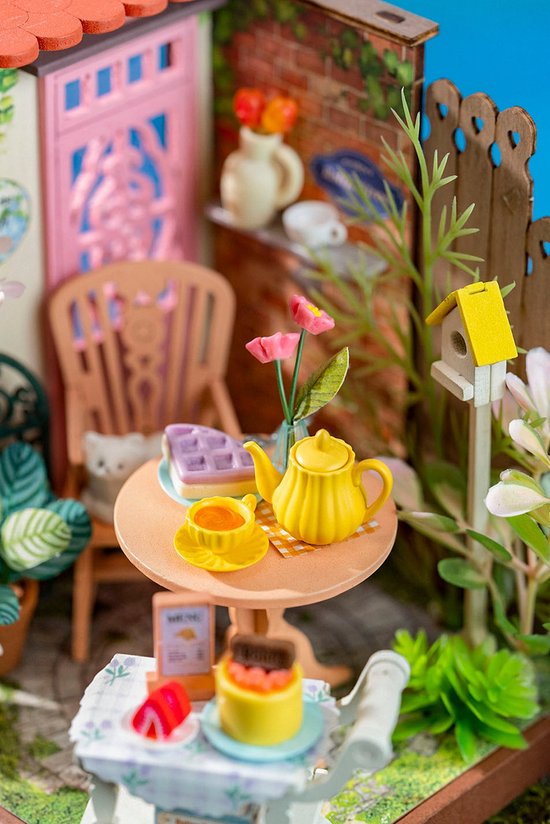 Miniatuur tuintafel met gele theepot, gebak, en bloemen. Een katje zit op de stoel.