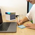 Een man met een laptop schrijft op een geel notitieblokje naast een witte mini-aircooler met kleurrijke LED-verlichting en een glas water.