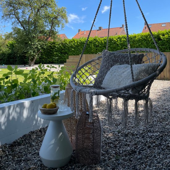 Grijze macramé hangstoel met diverse kussens in een tuinsetting.