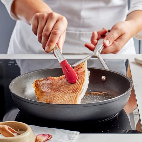 Chef-kok met rode tangen legt een stuk vis in een zwarte koekenpan op een inductiekookplaat.