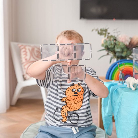 Een jongen met een gestreept shirt en blauwe shorts houdt Connetix-tegels voor zijn gezicht, kijkend naar de camera.