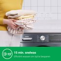 Une femme tenant une pile de vêtements tricotés, partiellement au-dessus d'un lave-linge gris foncé. Le panneau du lave-linge affiche le programme de lavage rapide de 15 minutes.