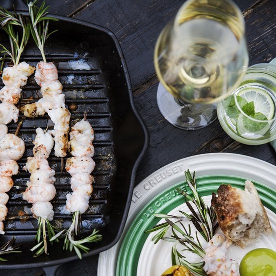 Gegrilde garnalen met rozemarijn in een zwarte grillpan, naast een glas witte wijn en een bord met brood.
