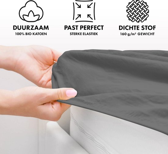 Een persoon toont de stofkwaliteit van een donkergrijs hoeslaken. Duurzaam, 100% bio katoen, sterk elastiek, dichte stof.