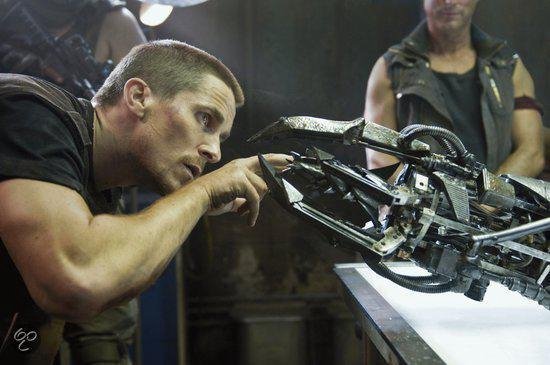 Christian Bale onderzoekt een robotarm, met een andere man op de achtergrond.