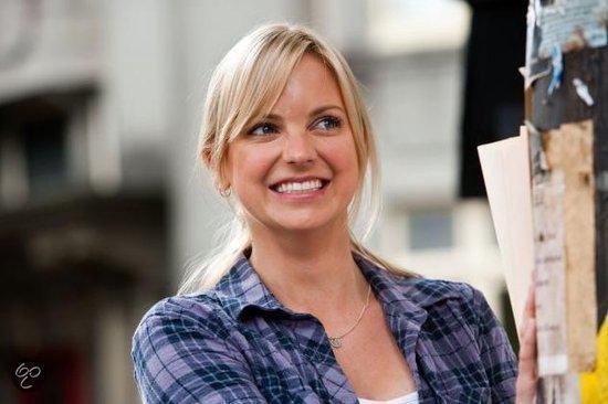 Een glimlachende blonde vrouw met een paarse ruitjesblouse en een zilveren ketting.