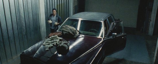 Man in grijze jas bij een auto met geld op de motorkap in een garage.