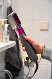 Een hand houdt een grijze haarstyler met roze accenten vast in een badkameromgeving.