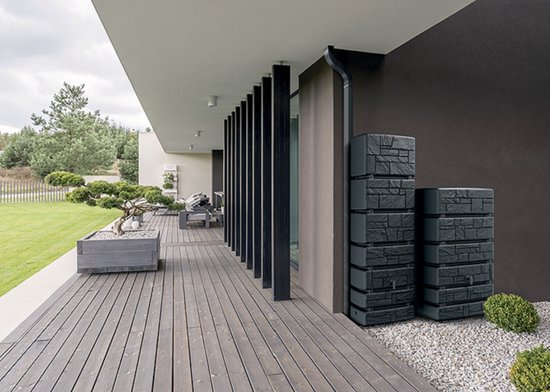 Twee antracietkleurige stapelbare regentonnen met steeneffect, naast een moderne bruine muur en houten terras.
