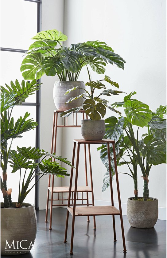 Drie kunstplanten op roestkleurige plantenstandaards in een woonkameropstelling.