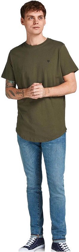 Man met tatoeages op zijn arm draagt een olijfgroene t-shirt met korte mouwen en ronde hals, met een klein logo op de borst, staand.