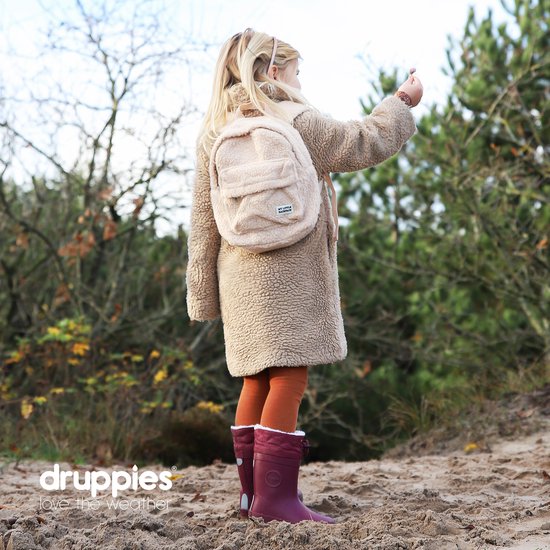 Meisje met blonde staart, teddy jas en paarse laarzen, staand op zand met bos op achtergrond.