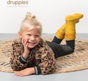 Een lachend blond meisje in luipaardprint jas ligt op een kleed met gele regenlaarzen aan.