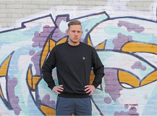 Man met handen in de zij draagt een zwarte Donnay trui met logo.