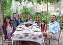 Een familie van acht personen, waaronder kinderen, zit aan een tafel in een tuin voor een maaltijd.
