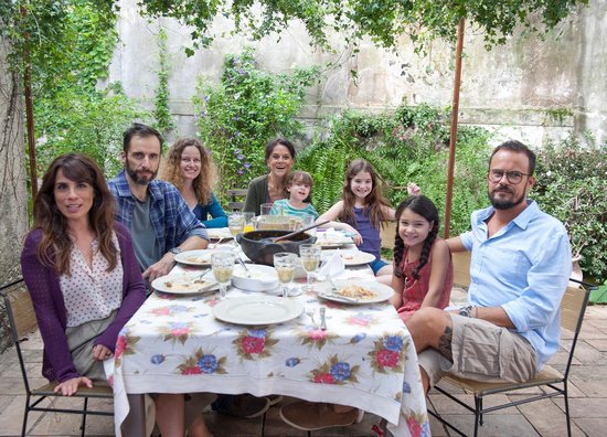 Een familie van acht personen, waaronder kinderen, zit aan een tafel in een tuin voor een maaltijd.