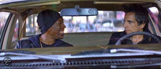 Twee mannen, één met een zwarte muts, zitten in een auto en kijken elkaar aan.