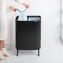 Une femme en robe blanche tricotée et bottines beiges ferme le sac poubelle bleu clair d'une poubelle Brabantia noire.