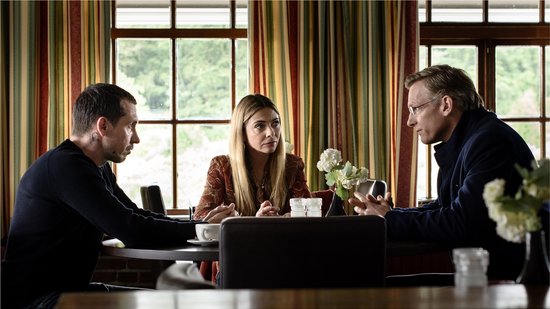 Drie personen zitten aan een tafel. Twee mannen en een vrouw. De vrouw draagt een oranje blouse.