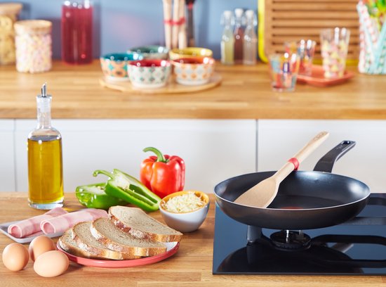 Een zwarte koekenpan met een houten spatel staat op een gasfornuis. Ingrediënten voor een ontbijt liggen ernaast.