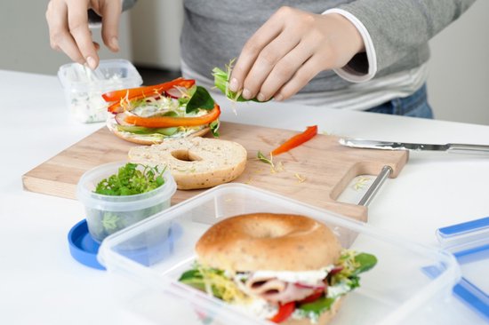 Une personne prépare un sandwich avec un bagel. Ingrédients et boîtes de conservation sur une planche à découper.