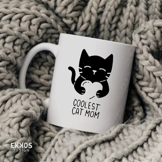 Witte mok met zwarte print van een kat die een hart vasthoudt met de tekst "COOLEST CAT MOM". Omringd door dik gebreide stof.