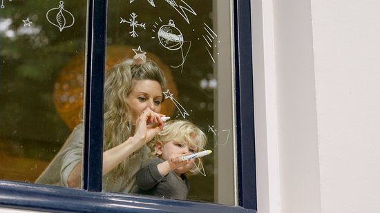 Vrouw met blond haar en een jong kind tekenen met witte stiften op een raam met kerstversiering.