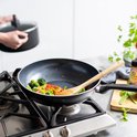Zwarte wokpan met groenten en houten spatel op gasfornuis. Handvat met "GREENPAN" logo.