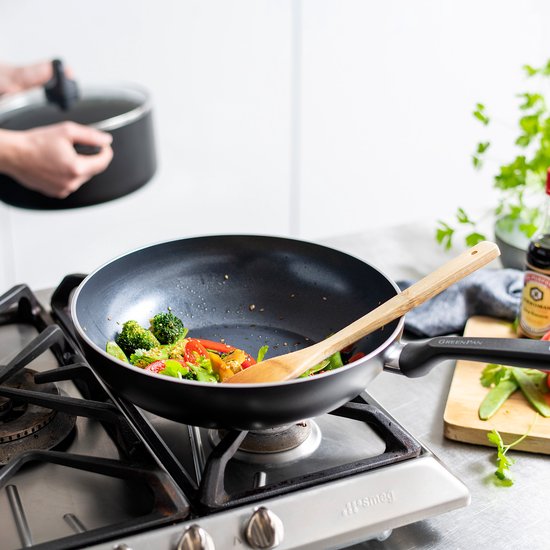 Zwarte wokpan met groenten en houten spatel op gasfornuis. Handvat met "GREENPAN" logo.