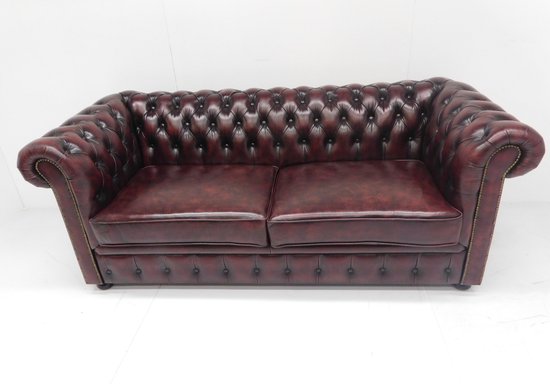 Klassieke bordeauxrode Chesterfield bank met diep gestoffeerde zitting en rugleuning, voorzien van knopen en goudkleurige decoratieve spijkers.
