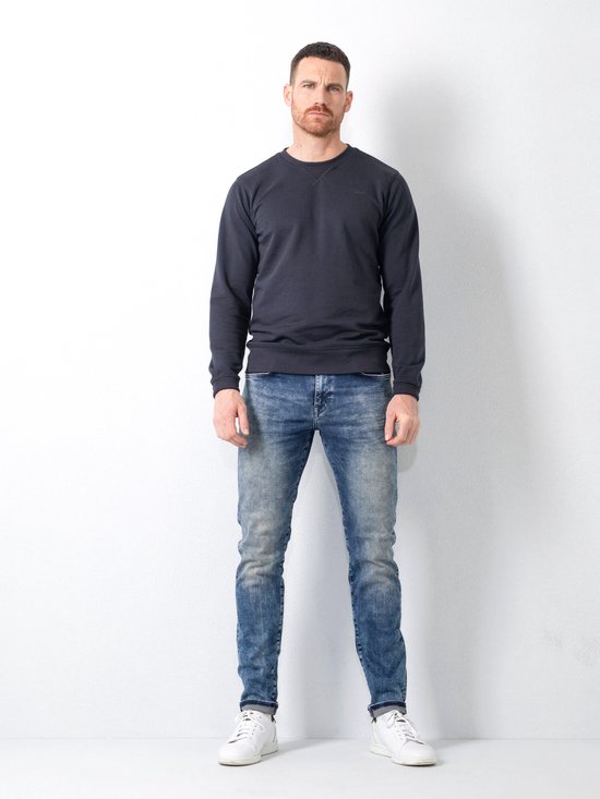Man met een blauwe slim fit jeans, wit shirt en witte sneakers, staand.