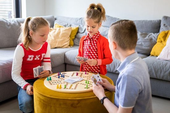 Drie kinderen spelen een bordspel aan de salontafel. De kinderen hebben pionnen en kaarten in hun handen. Het spel is een rond bord met gekleurde vakjes.
