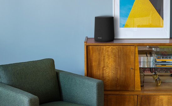 Zwarte Harman Kardon speaker op een houten kast naast een groene fauteuil.